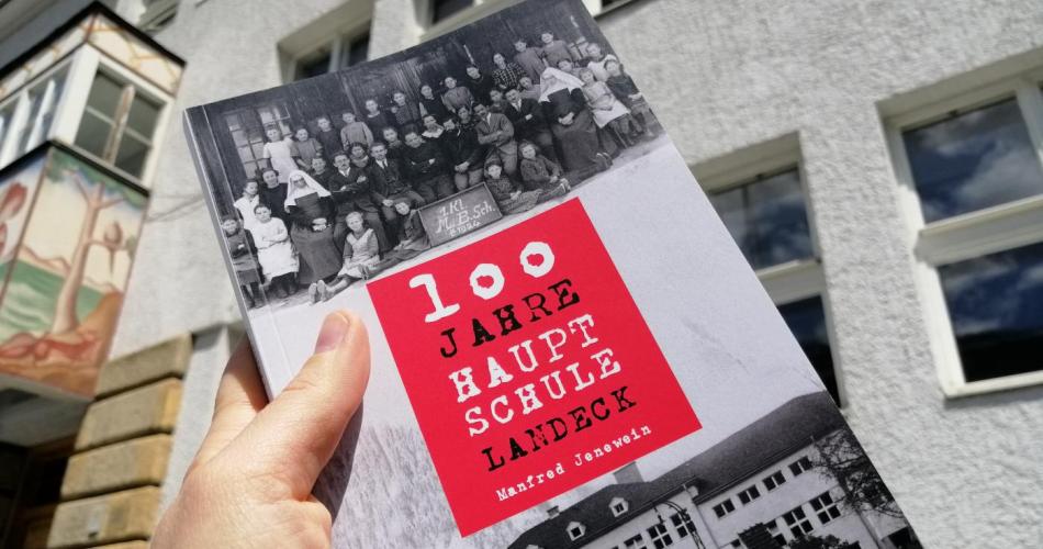Das Buch "100 Jahre Hauptschule Landeck" gehalten von einer Hand vor der Fassade der NMS Clemens Holzmeister